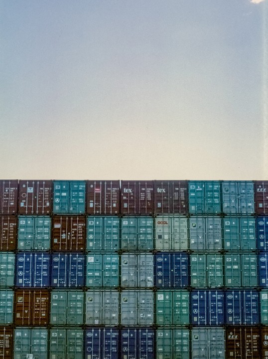 A wall of shipping containers in various colors is stacked under a clear sky.