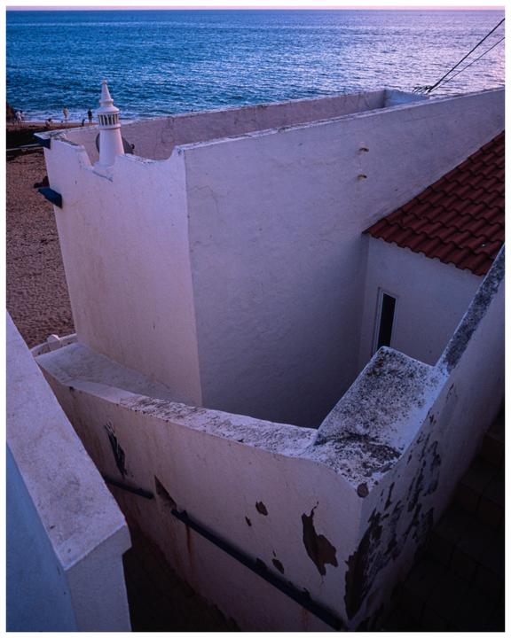 A white building with a red tile roof stands on a sandy beach next to the ocean.