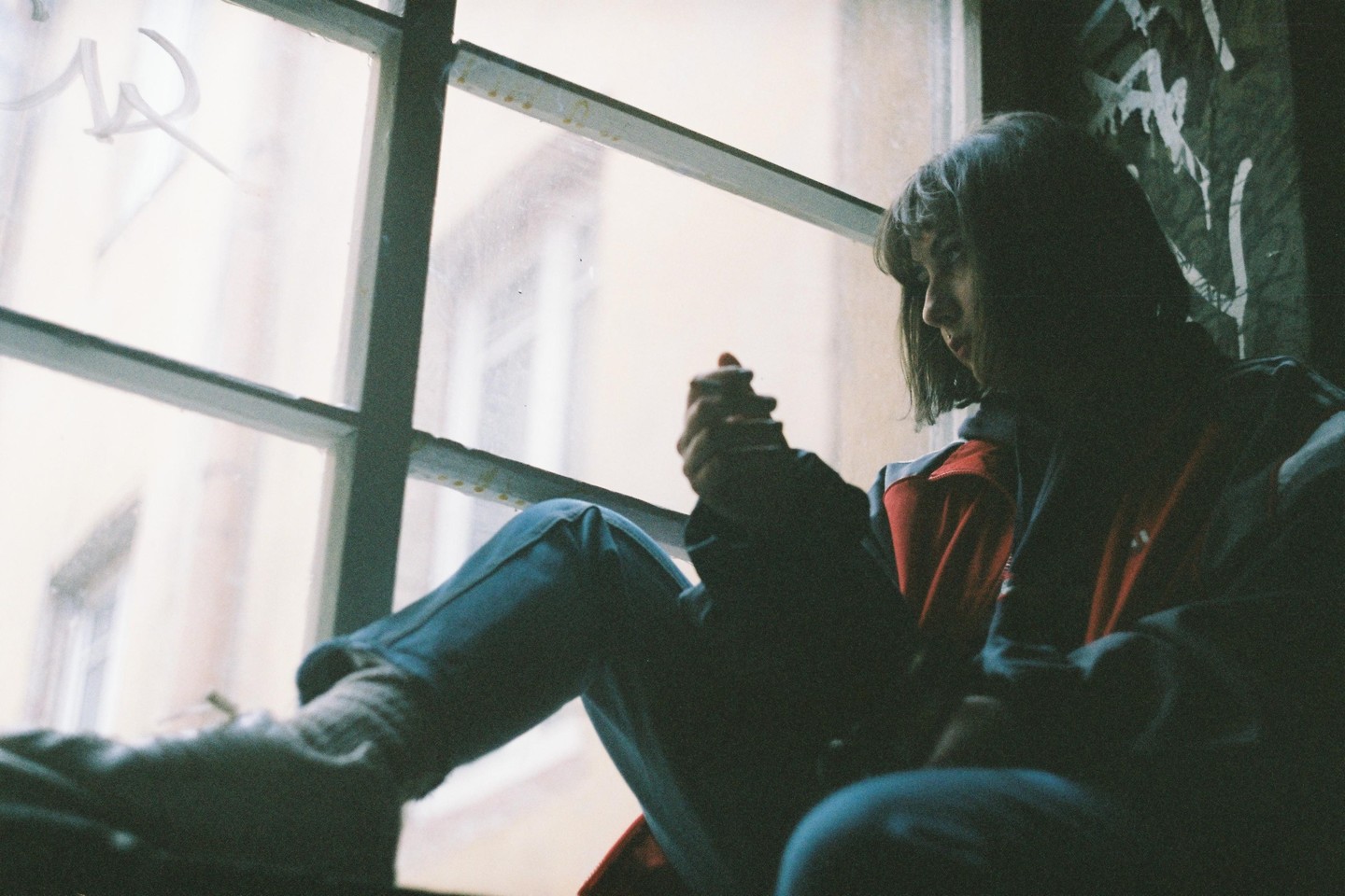 A woman sits on a window sill, looking out a window with graffiti on the wall next to her.