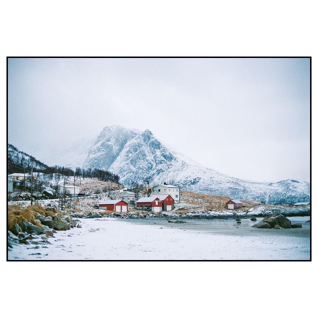 A small village with red and white houses sits at the base of a snow-covered mountain next to a frozen body of water.