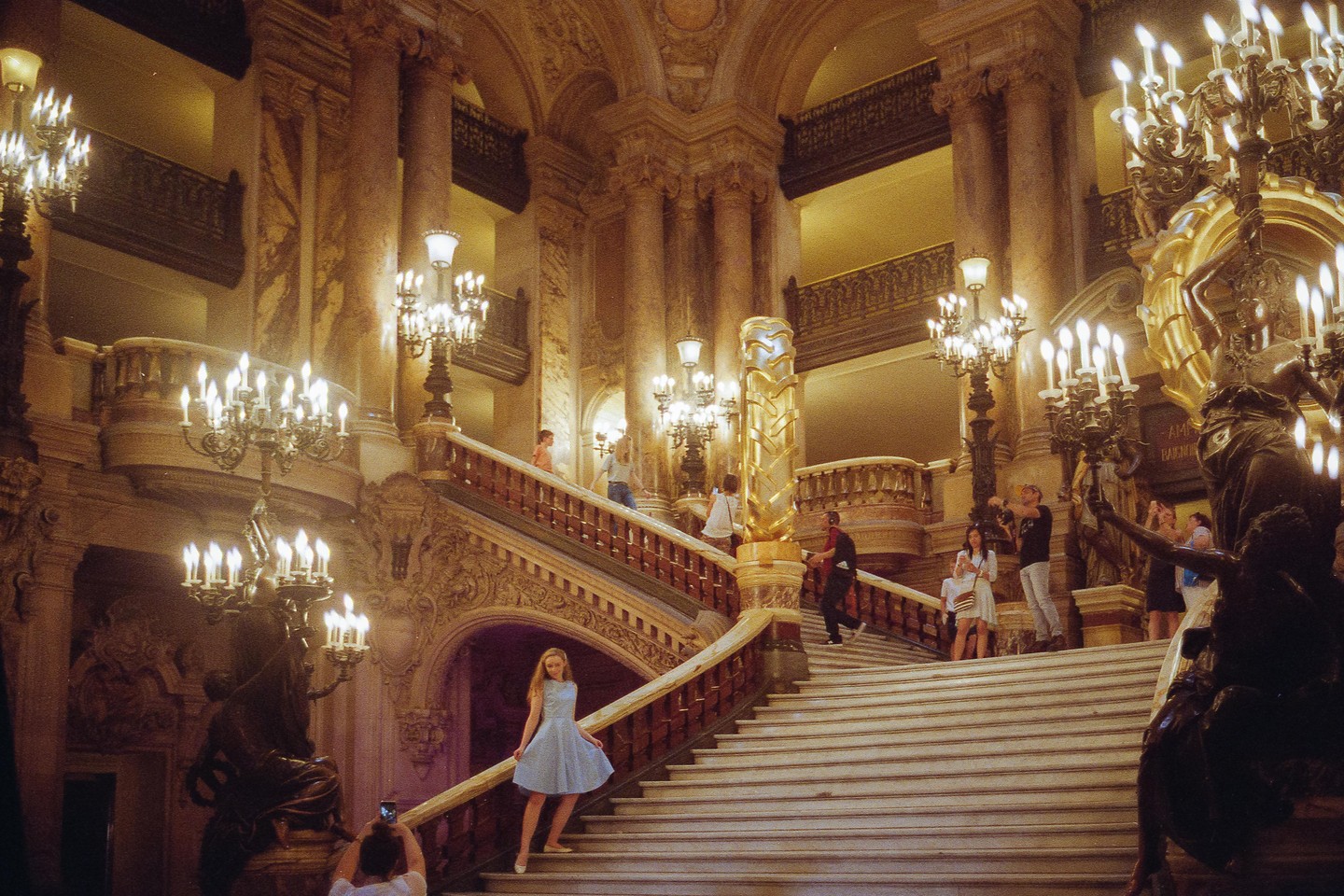 A woman in a light blue dress poses on a grand staircase in a richly decorated opera house, surrounded by ornate chandeliers and other people.
