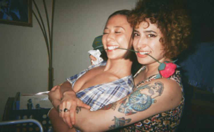 Two women are embracing and smiling with roses in their mouths.