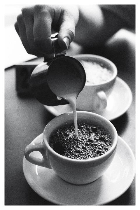 A hand pours milk into a cup of coffee, with another cup in the background.
