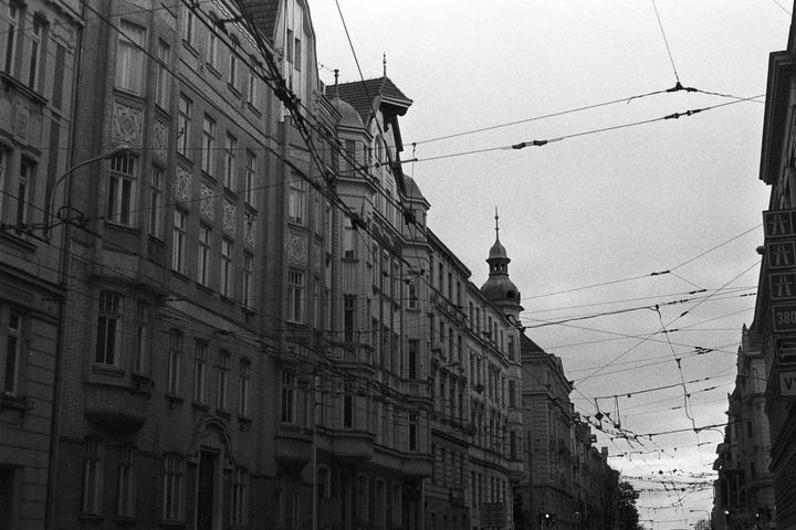 A street is lined with tall, ornate buildings and crisscrossing overhead wires under a cloudy sky.