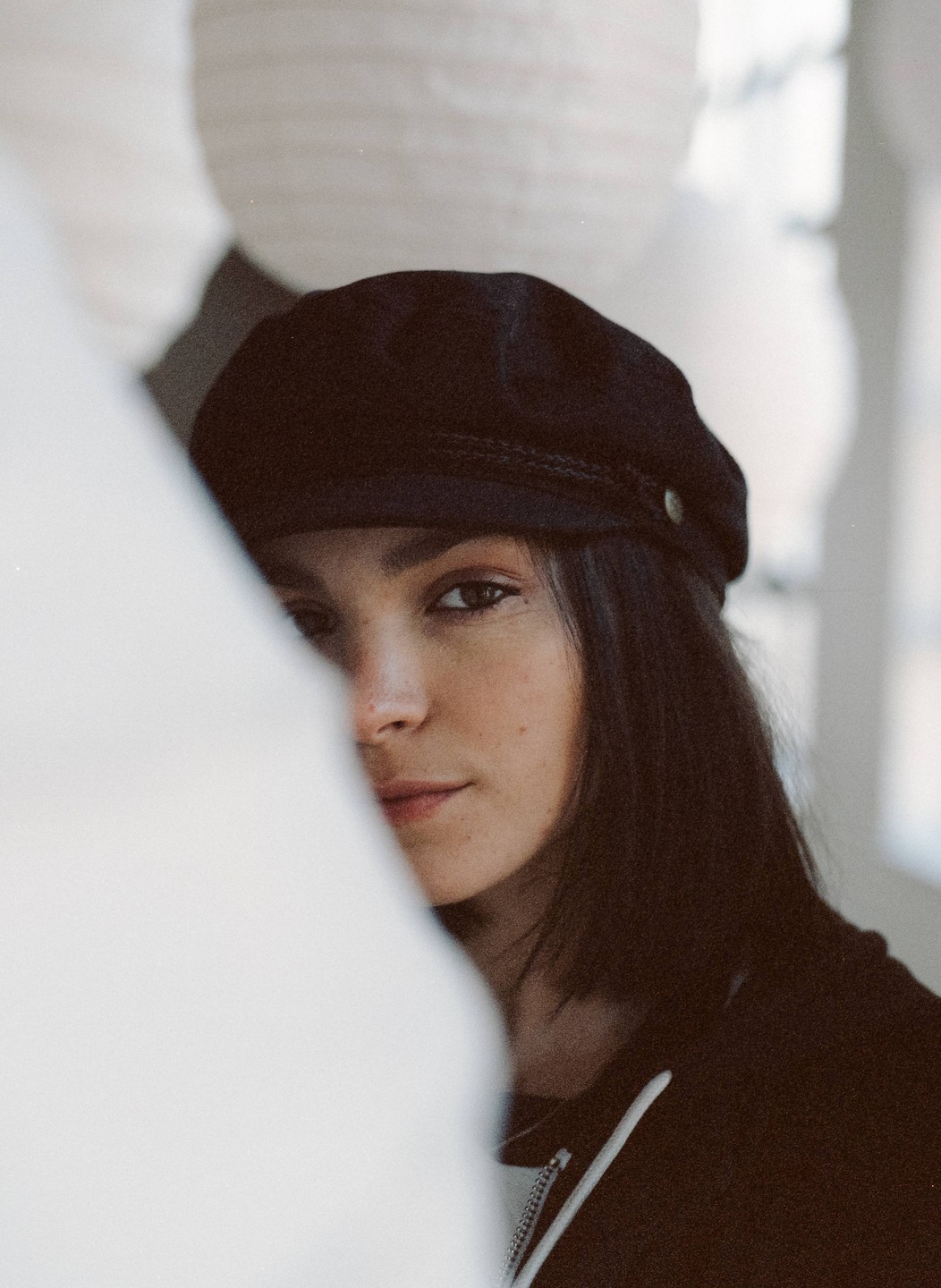 A woman wearing a black cap peeks from behind a white curtain.