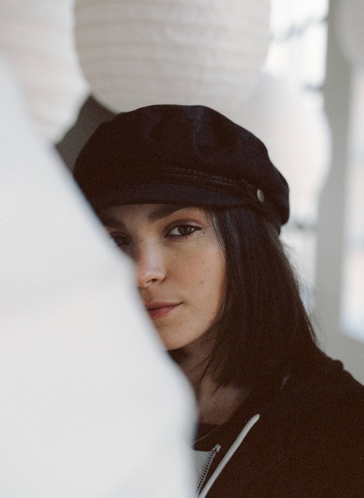 A woman wearing a black cap peeks from behind a white curtain.
