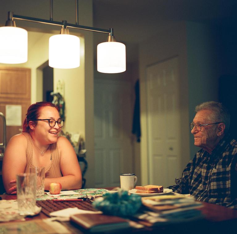 A woman and an elderly man sit at a table with food and drinks under hanging lights.