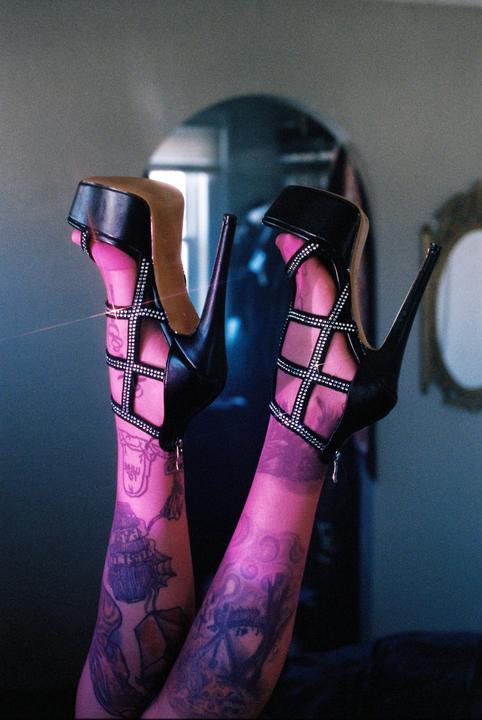 A person's legs with tattoos and pink stockings are visible, wearing black high heels with studded straps.
