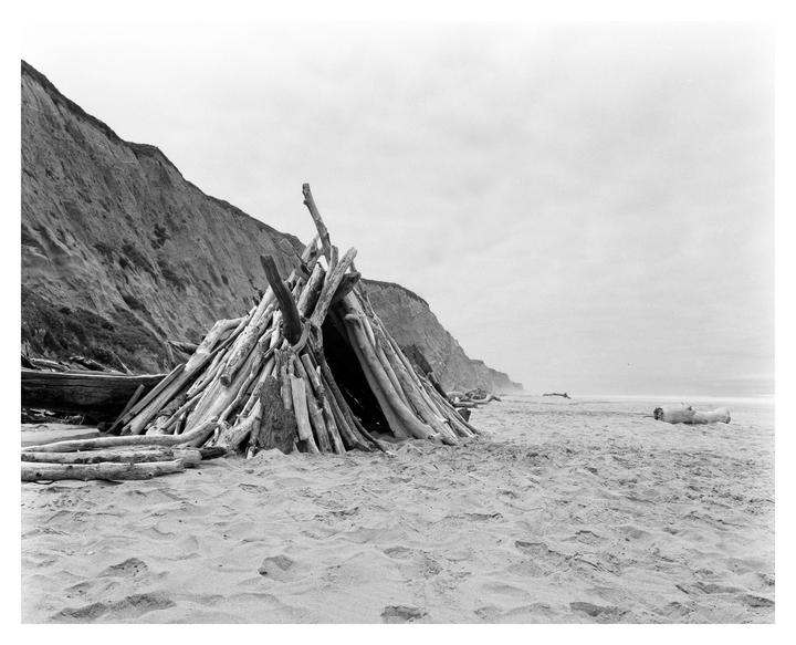 A driftwood hut stands on a sandy beach next to a large cliff under a cloudy sky.
