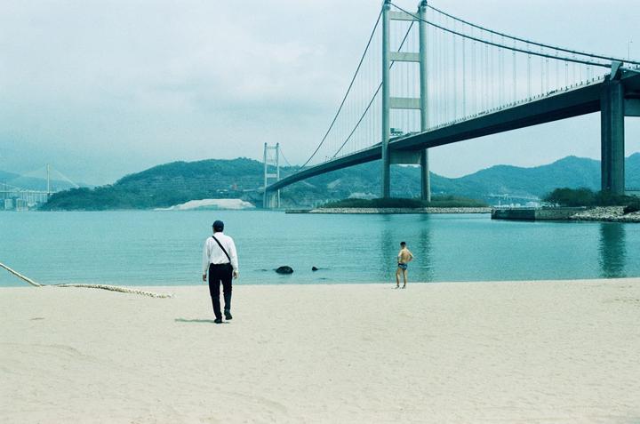 Two men stand on a sandy beach with a large suspension bridge and mountains in the background.