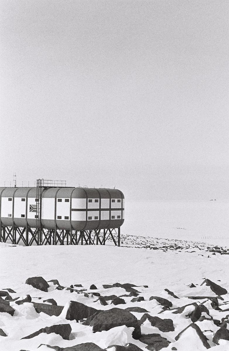 A research station stands on stilts in a snowy, rocky landscape under a bright sky.