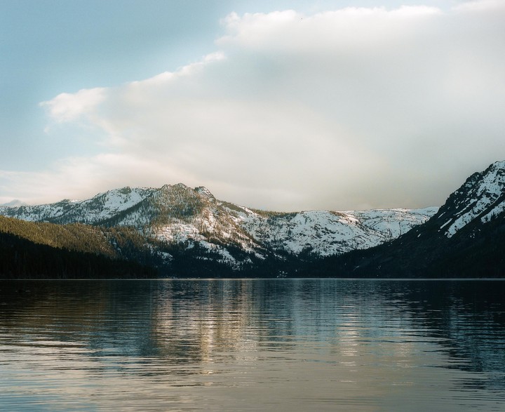 A body of water reflects snow-covered mountains under a cloudy sky.