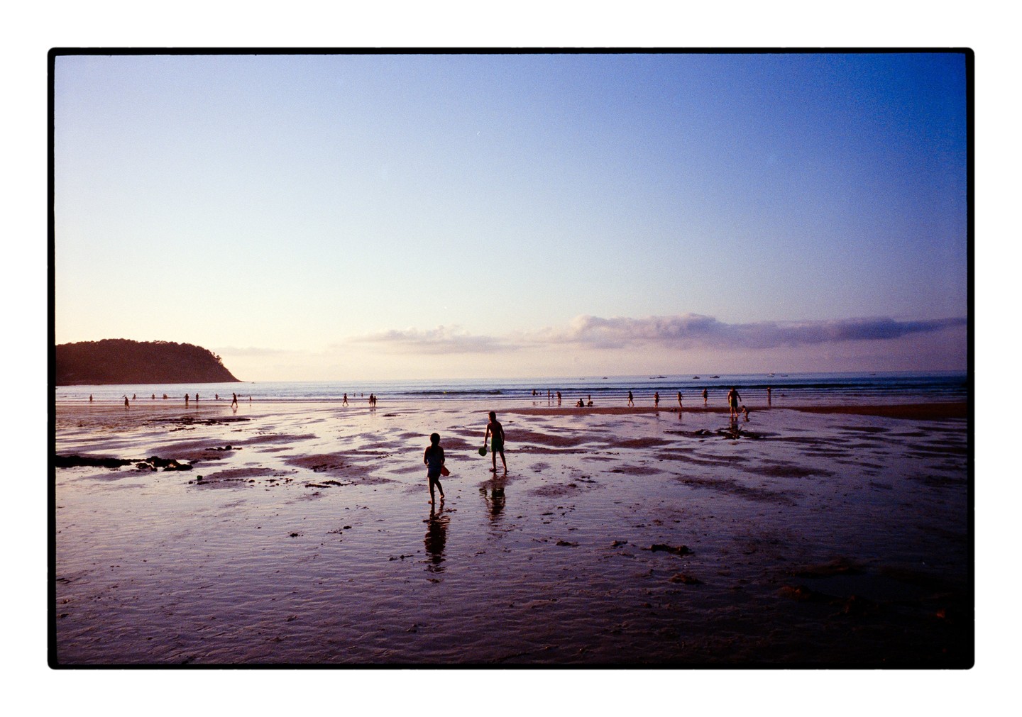 Two children play on a wet beach at dusk with many other people in the distance and a large hill on the left.