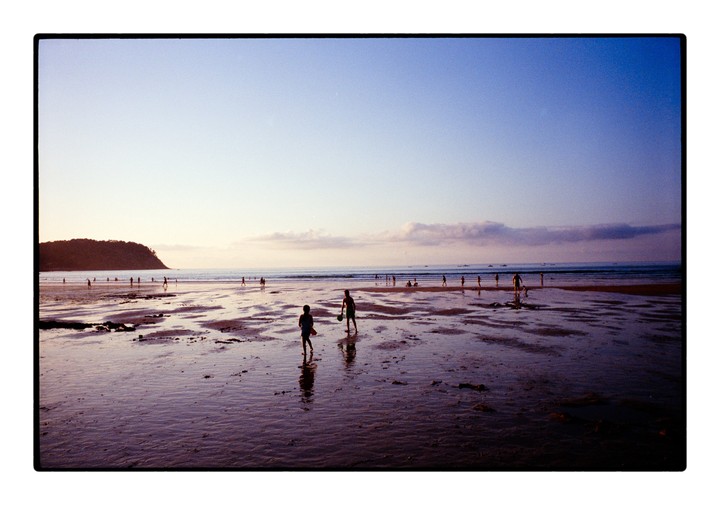 Two children play on a wet beach at dusk with many other people in the distance and a large hill on the left.