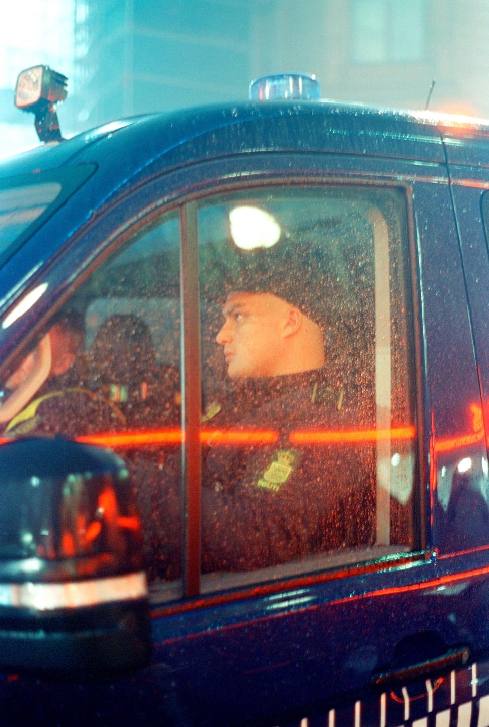 A man in uniform looks out the window of a police vehicle at night.
