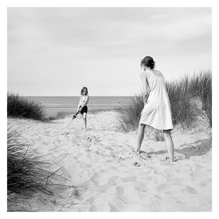 A woman and a girl play cricket on a sandy beach with tall grass.