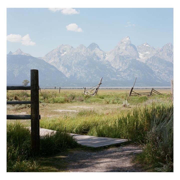 A wooden fence and boardwalk lead to a grassy field with mountains in the background.
