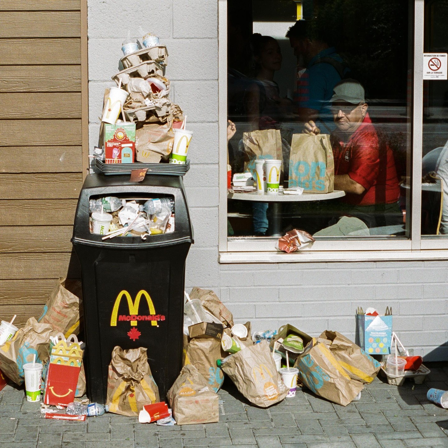 A McDonald's trash can overflows with garbage next to a window where people are eating.
