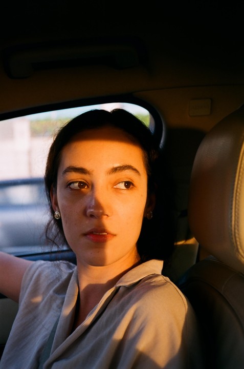 A woman with dark hair looks out of a car window as golden light illuminates half of her face.