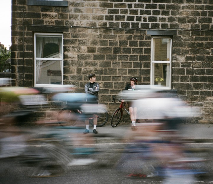 Two men stand with their bicycles on a sidewalk next to a brick building as blurred cyclists speed past on the street.