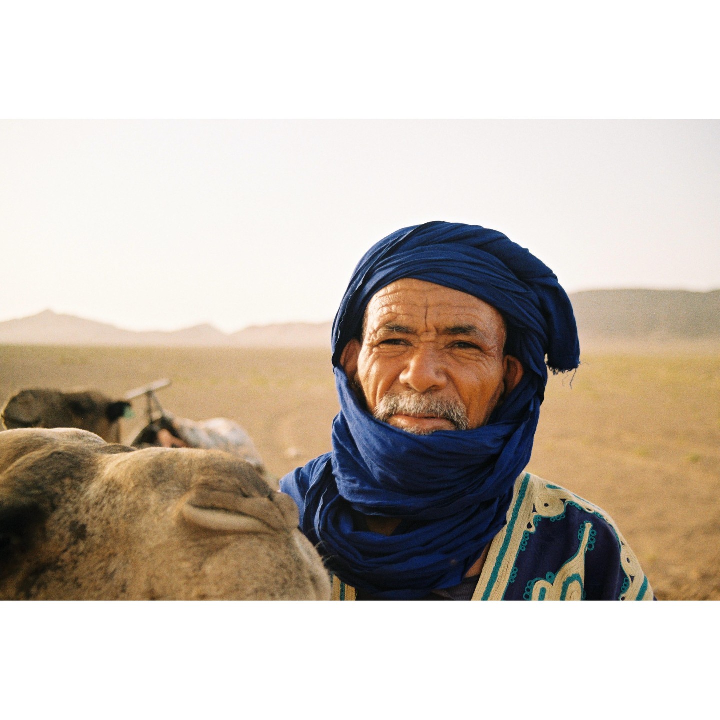 A man wearing a blue turban and patterned robe stands next to a camel in a desert landscape.