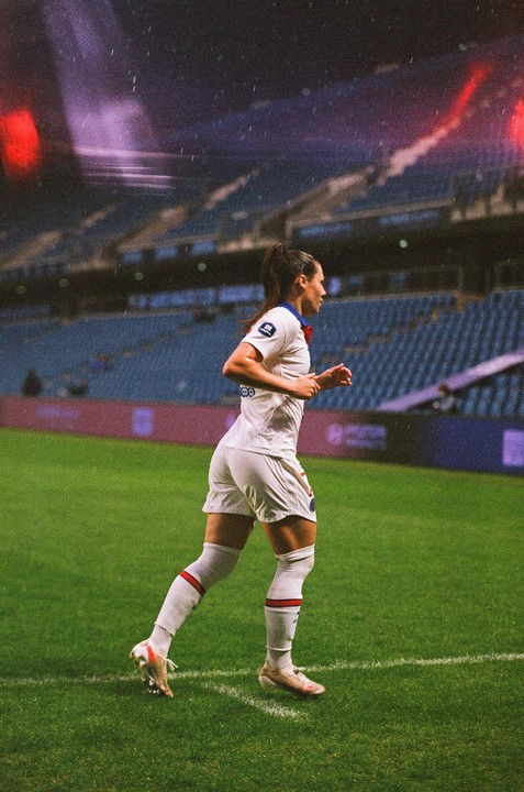 A woman in a white uniform runs across a green soccer field at night.