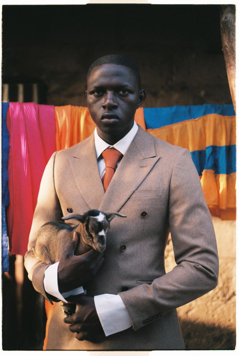 A man in a suit holds a baby goat in front of colorful fabric.