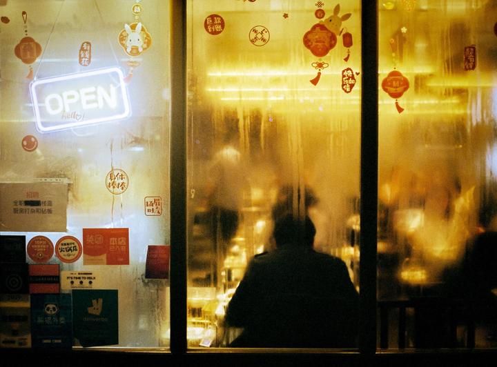 A person sits inside a restaurant with a neon "OPEN" sign and various stickers on the window.