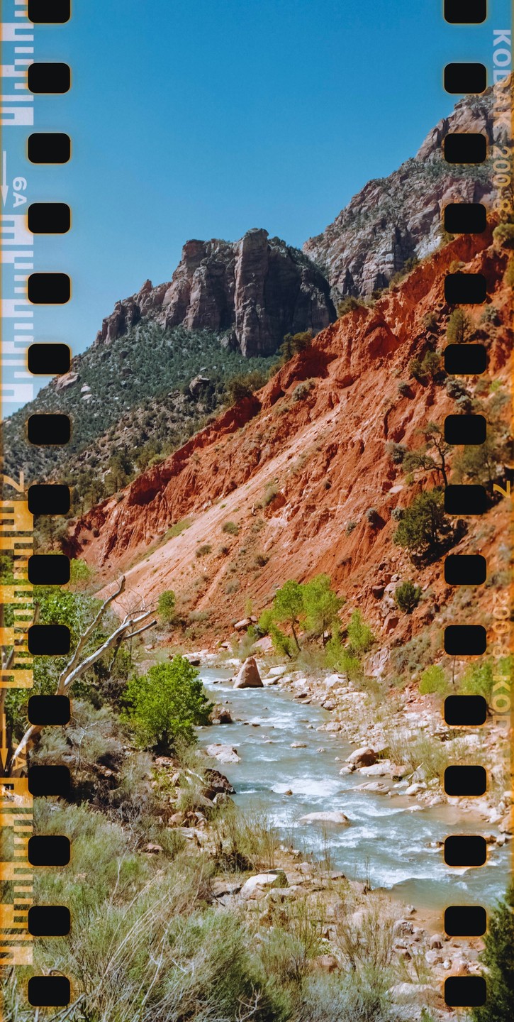 A river flows through a valley with steep red rock cliffs and green vegetation under a clear blue sky.