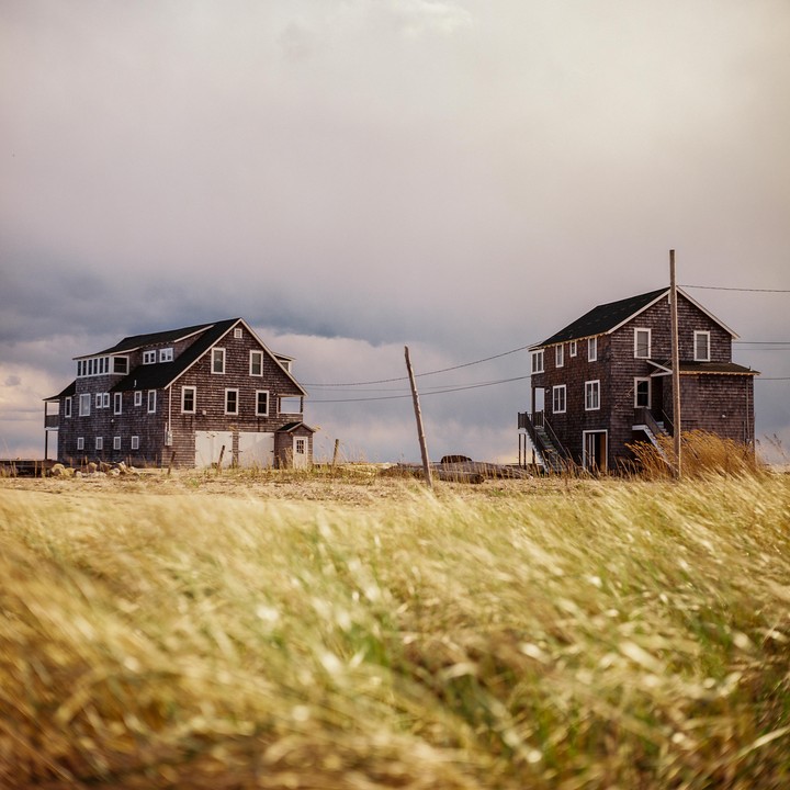 Two weathered wooden houses stand on a grassy dune under a cloudy sky.