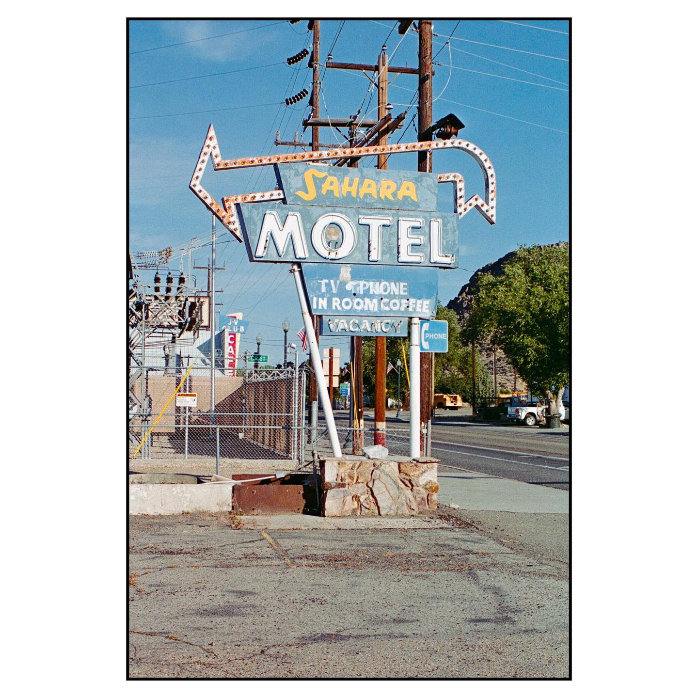 A weathered Sahara Motel sign with an arrow pointing left stands beside a road with power lines overhead.