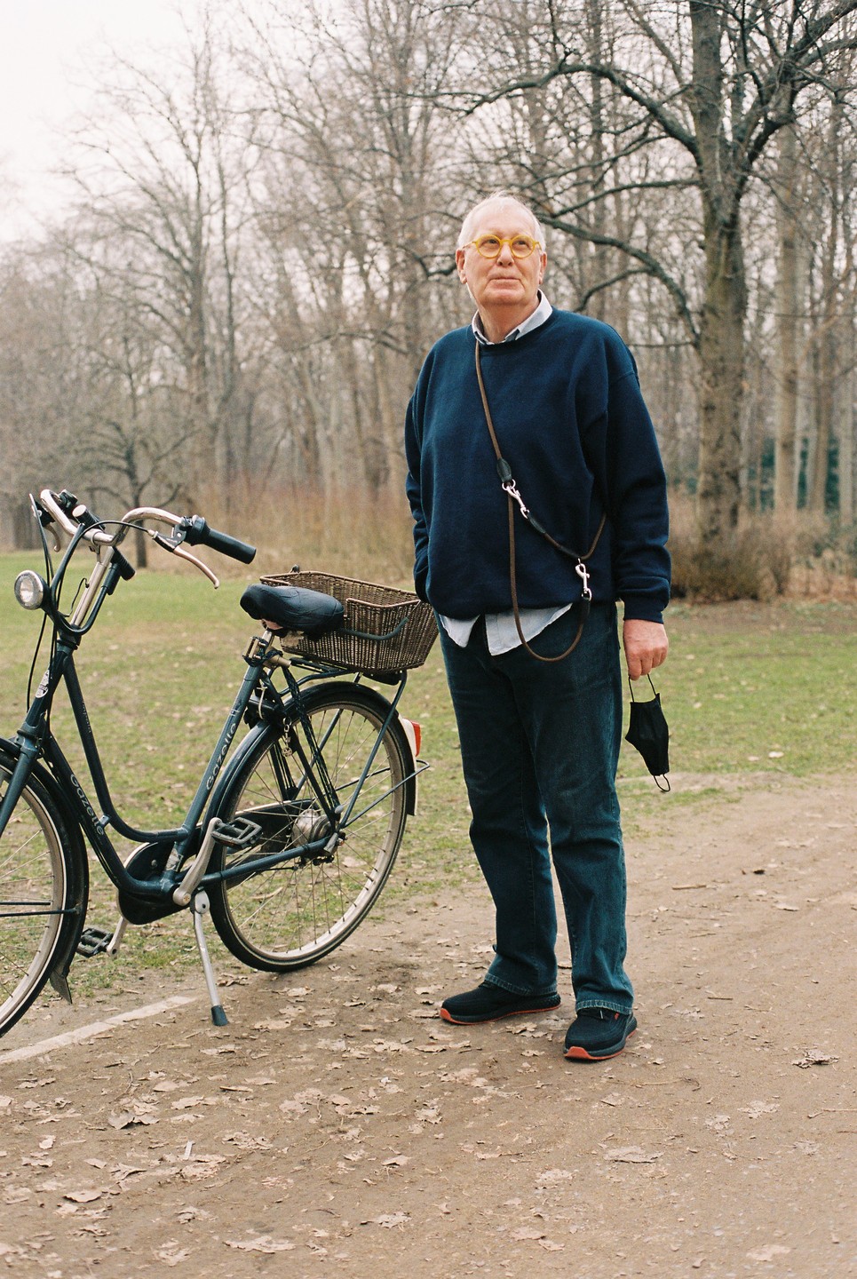 A man stands next to a bicycle on a dirt path with trees in the background.