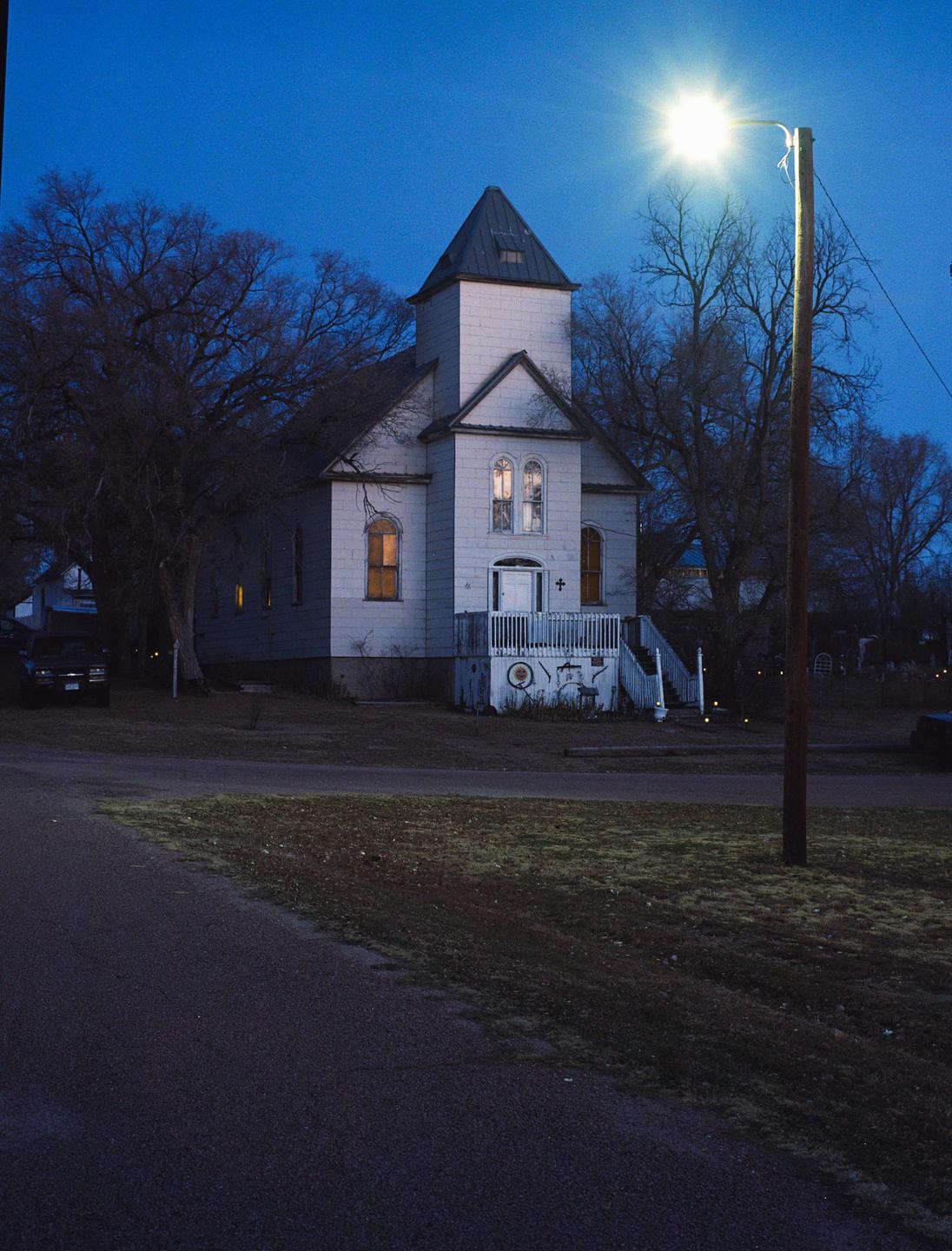 A white church with a dark roof and steeple stands among bare trees under a deep blue sky at night, illuminated by a bright street light.