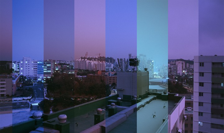 A cityscape with buildings and trees is visible from a rooftop, with the sky showing bands of different colors.