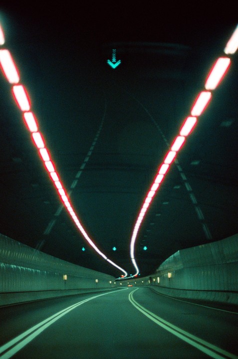 A long exposure shot shows the interior of a tunnel with red lights curving along the ceiling and a green arrow sign.