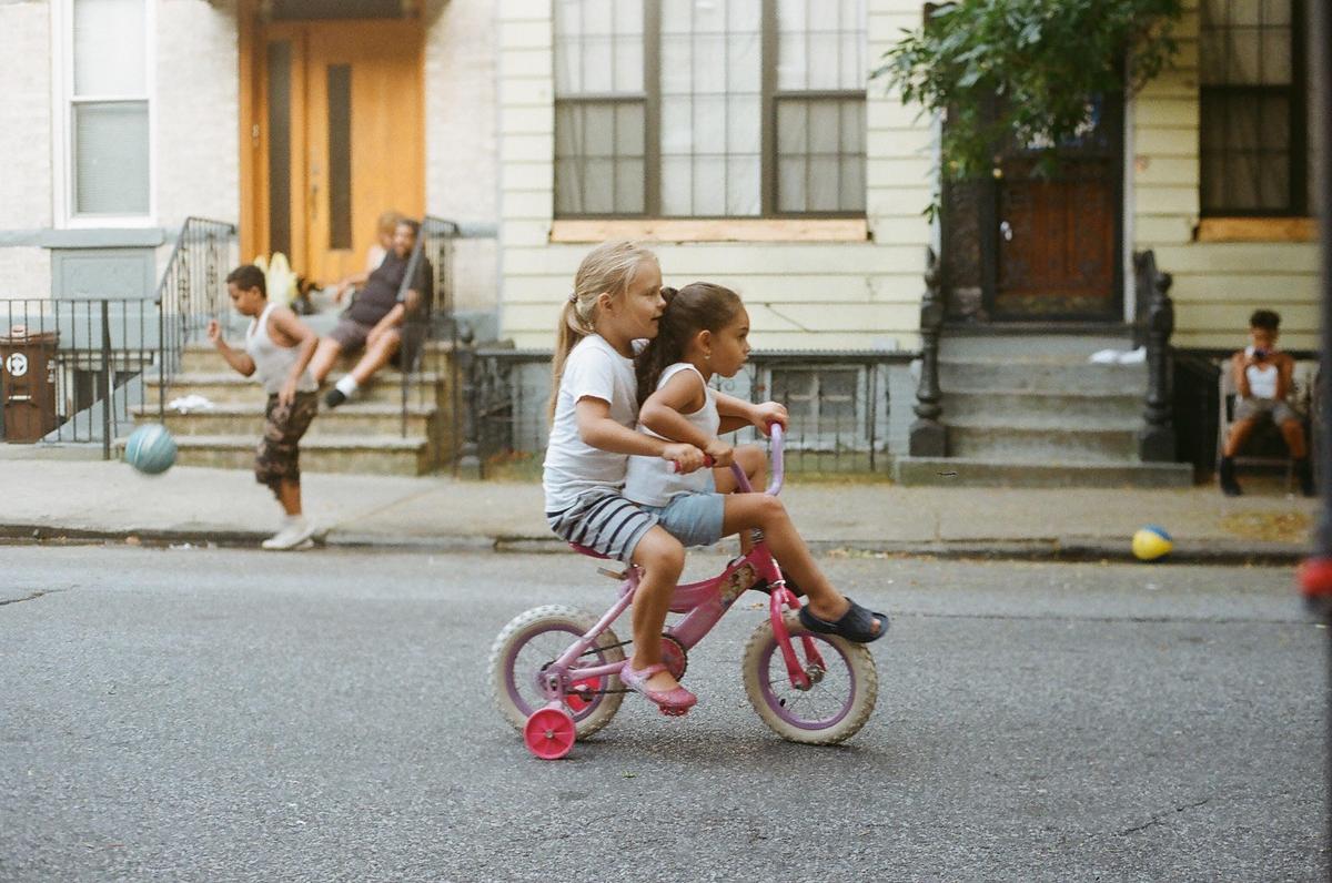 Two girls ride a pink bicycle on a street while other children play and sit on stoops in the background.