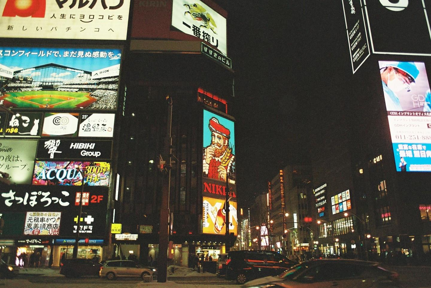 A street at night is illuminated by numerous bright billboards and signs, with cars and buildings visible.