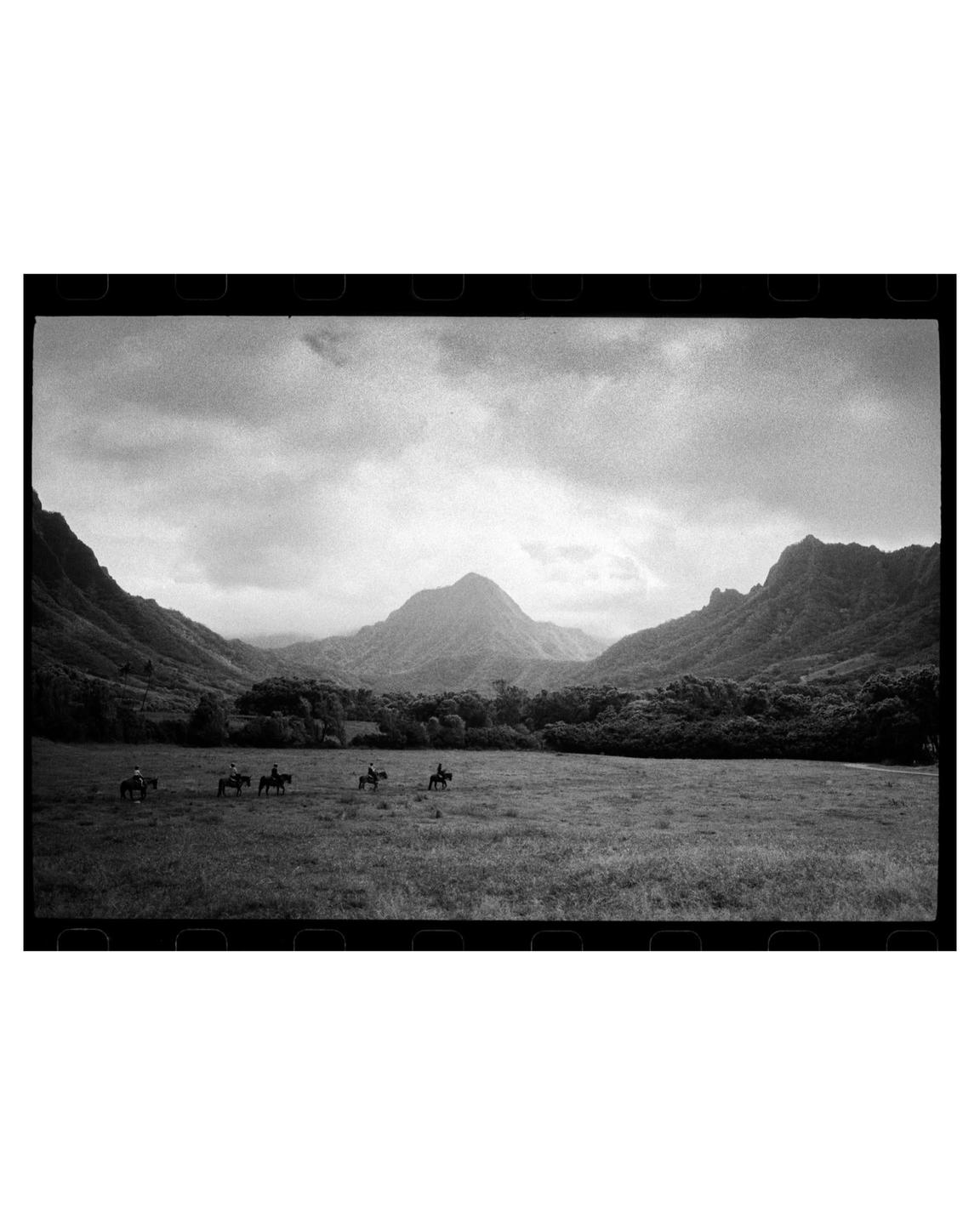 A group of people on horseback ride through a grassy field with mountains in the background under a cloudy sky.