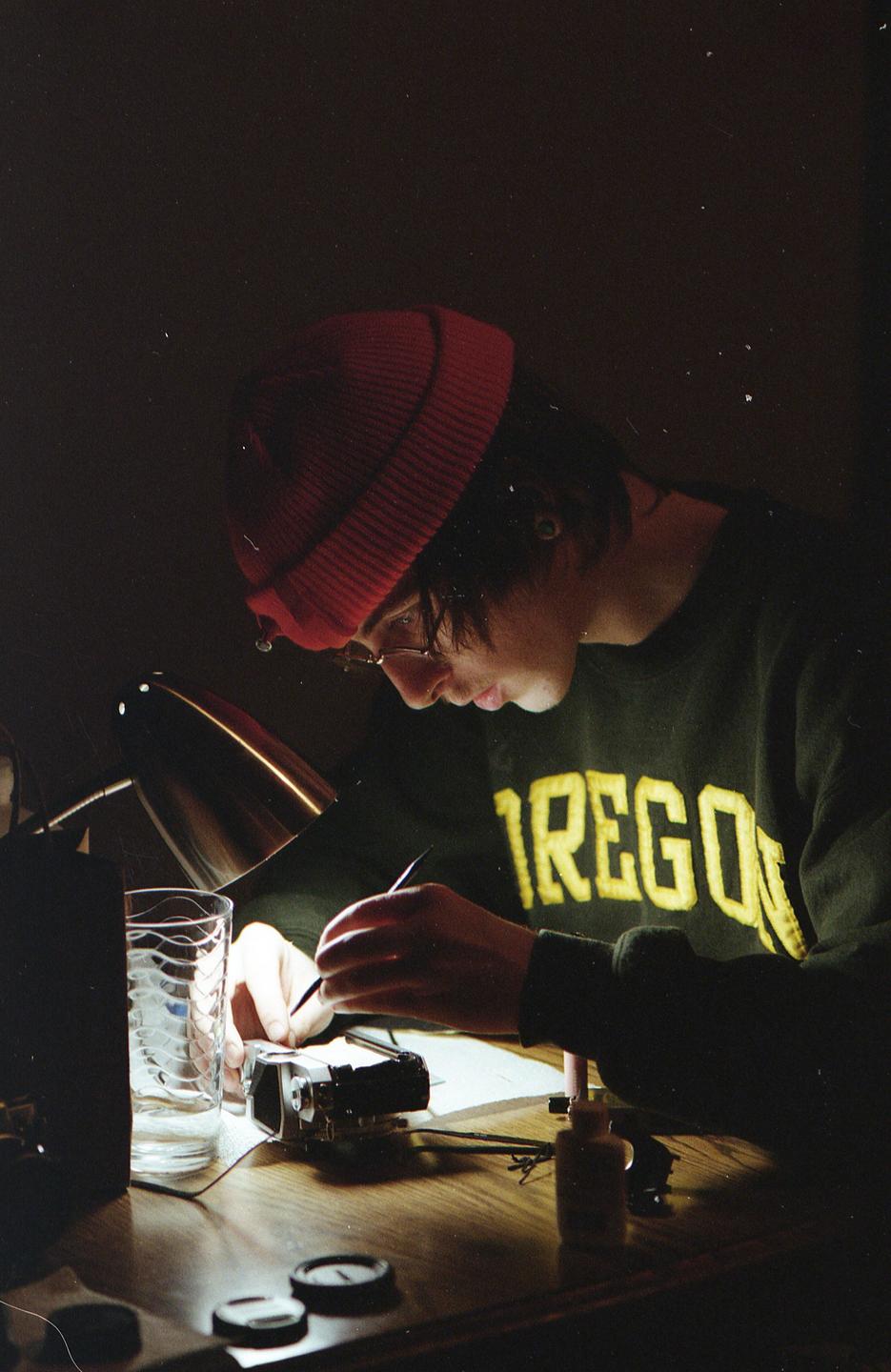 A person wearing a red beanie and a green sweatshirt with "OREGON" written on it is working on a camera at a wooden desk, illuminated by a desk lamp.