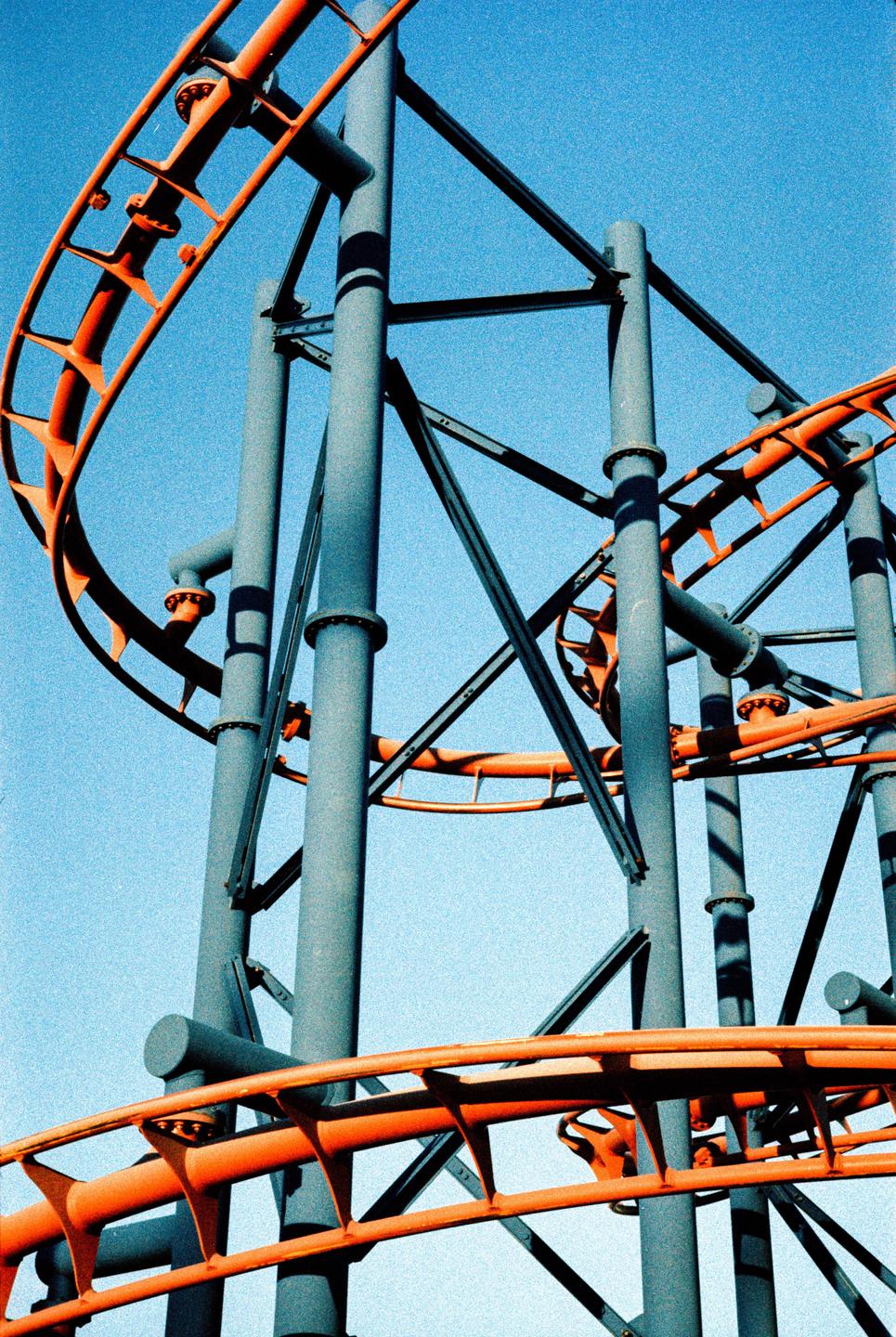 A roller coaster with orange tracks and grey supports stands against a clear blue sky.