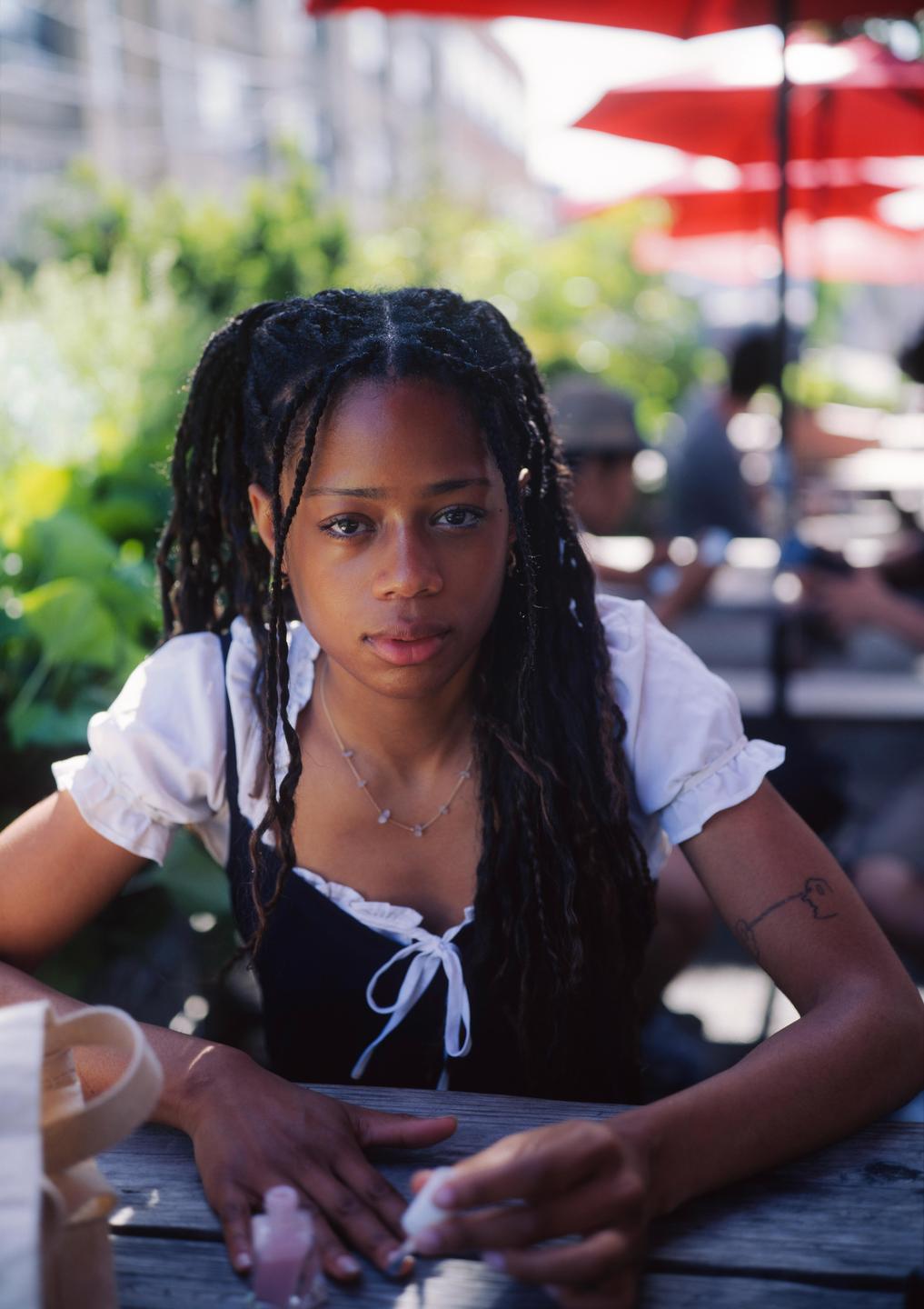 A woman with dreadlocks sits at a wooden table, applying nail polish to her fingernails.