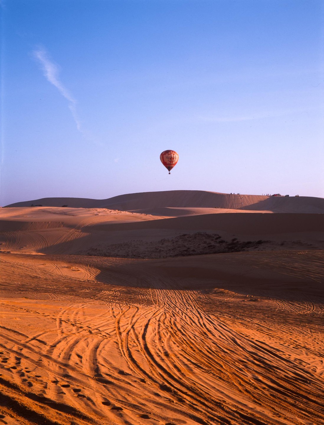 A hot air balloon floats above sand dunes with tire tracks and footprints.