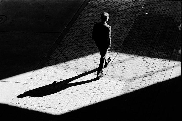 A man walks on a patterned sidewalk, casting a long shadow in a high-contrast black and white street scene.