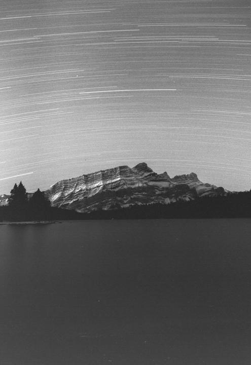 A long exposure photograph captures star trails above a mountain range and a lake.