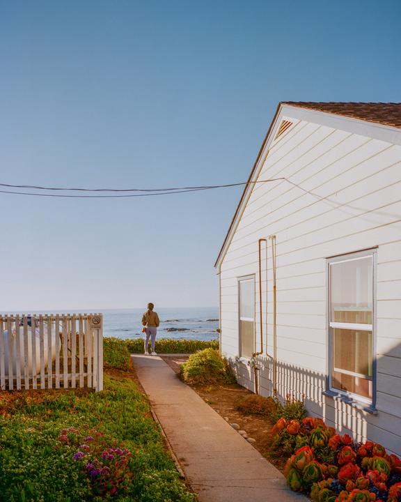 A person walks along a path towards the ocean next to a white house with a picket fence.
