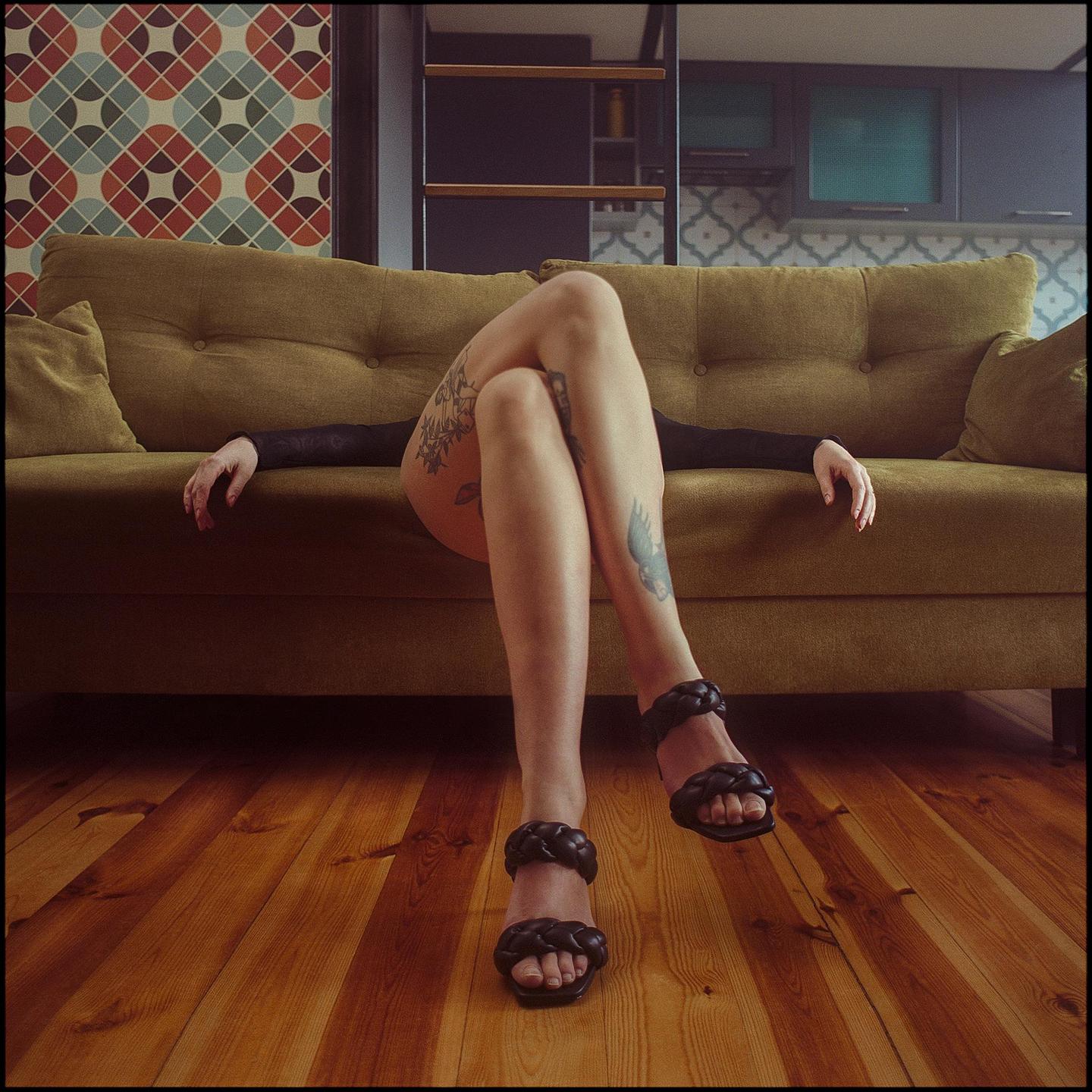 A woman with tattooed legs and black sandals is lying on a green sofa with her arms outstretched.