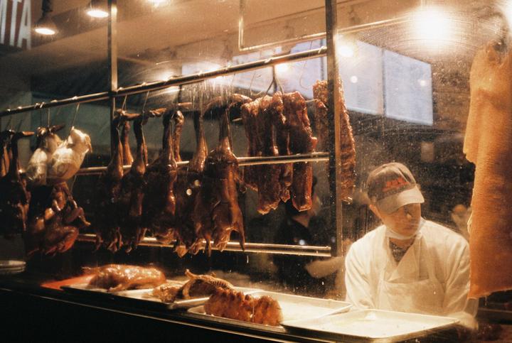 A man in a white uniform and cap stands behind a glass display case filled with hanging roasted ducks and other meat.
