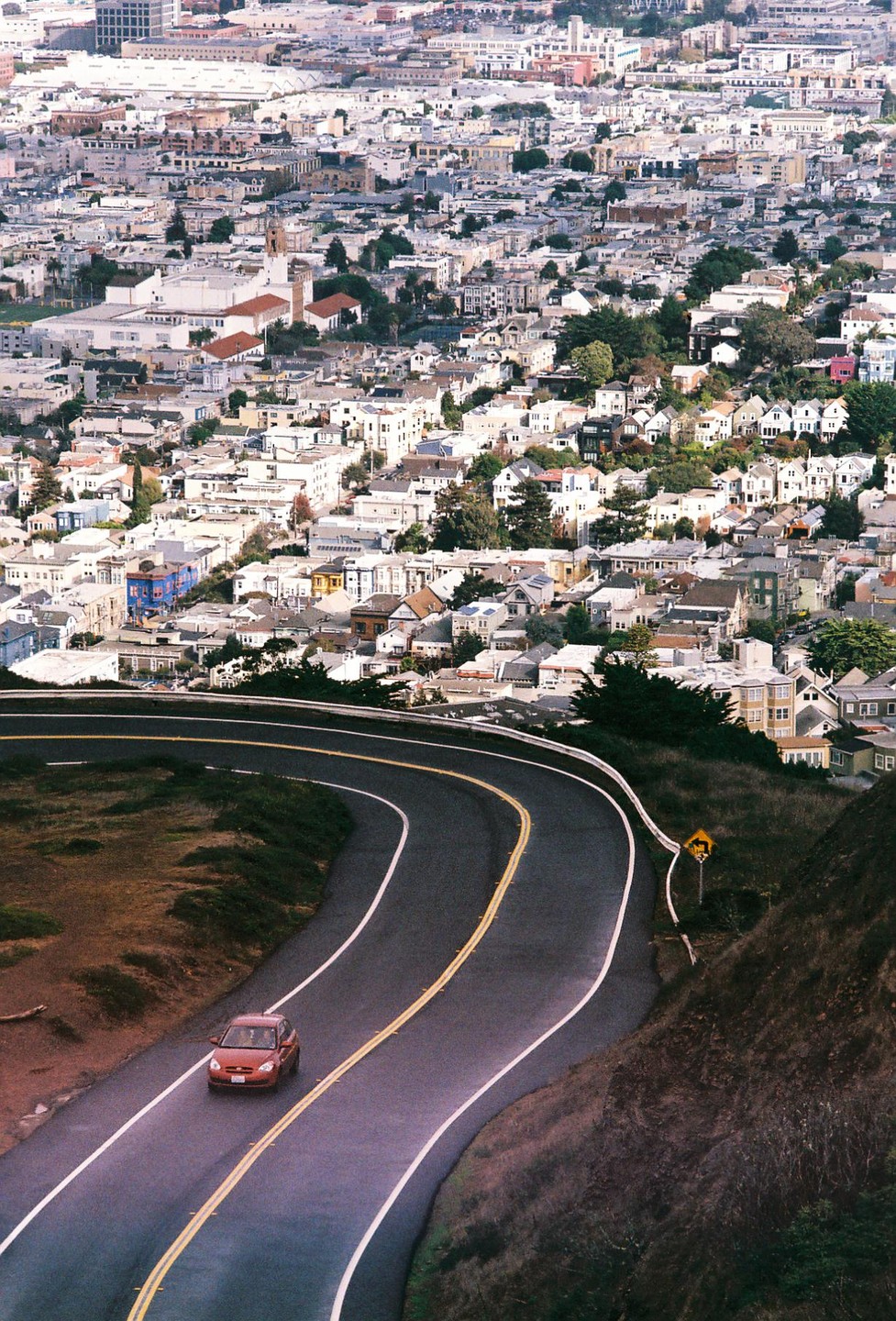 A red car drives on a winding road overlooking a city.