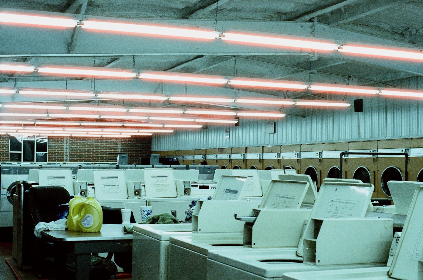 A laundromat interior is illuminated by rows of fluorescent lights, with washing machines and dryers lining the walls.