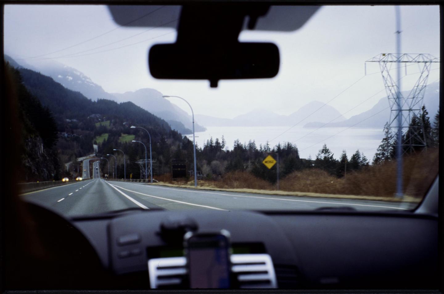 A view from inside a car shows a highway winding through mountains alongside a body of water.